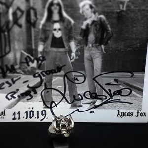 May include: A black and white photo of a band signed by the band members. The photo is signed "We are the Ring - Growler" and "Lucas Fox". The date "21.10.19" is written below the signatures. A silver ring with a detailed skull design is in the foreground.