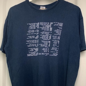 May include: A dark navy blue short-sleeved t-shirt featuring a white graphic print on the chest. The print displays a list of computer command line instructions and technical terminology arranged in a grid format, oriented upside down.