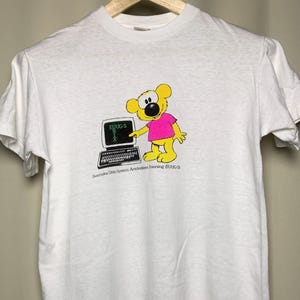 Könnte beinhalten: Ein weißes Kurzarm-T-Shirt mit einer Grafik eines gelben Cartoon-Bären in einem pinken Oberteil, der neben einem schwarzen Laptop steht. Auf dem Bildschirm steht der Text EUUG-S UNIX und unter dem Laptop der Text Svenska Unix System Användares Förening (EUUG-S).