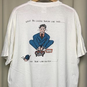 以下が含まれることがあります： 背面に漫画のグラフィックが描かれた白いTシャツ。グラフィックは、小さな車の上に座っている青いスーツを着た男性を描いており、「あなたはあなたの車をこのように選びますか...そしてあなたのLANスイッチ...？」というテキストが書かれています。