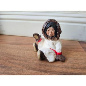 May include: A ceramic figurine of an Inuit child in a seated position. The child wears a white parka with a fur-lined hood and red accents. The figurine has a smiling face and is waving a hand.