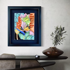 May include: A framed watercolor painting featuring a green frog peeking out from behind a tree trunk, surrounded by colorful tropical flowers and foliage. The artwork is set in a dark blue frame, adding a striking contrast to the vibrant colors of the painting.