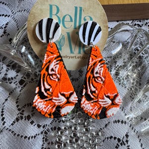 May include: A pair of handmade earrings featuring a tiger design. The earrings have a black and white striped circular top and a triangular orange, black, and white tiger face pendant. The earrings are displayed on a lace surface.