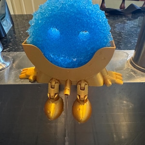 May include: A blue, textured sponge in a gold-colored holder with a whimsical design. The holder has a face-like appearance with two eye-shaped holes and two gold-colored legs. Water is flowing from the bottom of the holder.