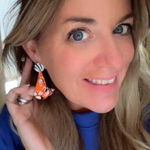 May include: A woman wearing a pair of orange and white dangle earrings. The earrings have a black and white striped top and a patterned orange bottom. The woman is wearing a blue shirt and a silver bracelet.