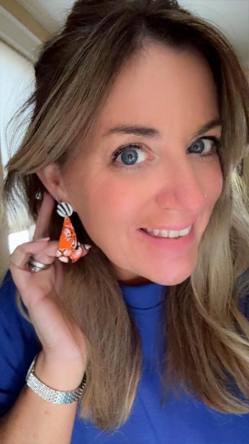 May include: A woman wearing a pair of orange and white dangle earrings. The earrings have a black and white striped top and a patterned orange bottom. The woman is wearing a blue shirt and a silver bracelet.