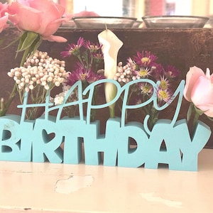 May include: A light blue wooden "HAPPY BIRTHDAY" sign with a heart-shaped cutout. The sign holds various flowers, including pink roses, white calla lilies, and purple and white wildflowers. The arrangement is set against a rustic wooden background.