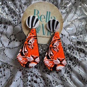 May include: A pair of handmade earrings featuring a tiger design. The earrings have orange, black, and white details. The top of the earrings are white circles with black stripes. The earrings are displayed on a round tag with the text "Bella Vie" and "jewelryart.etsy.com".