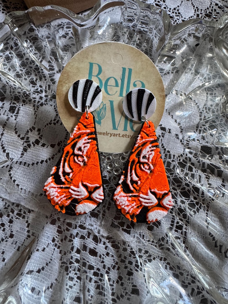 May include: A pair of handmade earrings featuring a tiger design. The earrings have orange, black, and white details. The top of the earrings are white circles with black stripes. The earrings are displayed on a round tag with the text "Bella Vie" and "jewelryart.etsy.com".