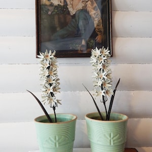 Puede incluir: Dos macetas de cerámica verde con flores de metal blanco. Las macetas tienen un diseño texturizado y están sobre una superficie de madera.