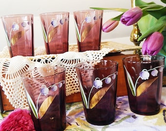 Könnte beinhalten: Sechs violette Glasbecher mit goldenen Akzenten und einem weißen Blumendesign. Die Becher sind auf einer weißen Häkelspitze angeordnet.