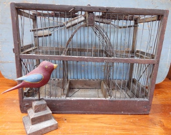 May include: A vintage wooden birdcage with a wire mesh interior. The cage is brown and has a distressed finish. A small, painted wooden bird is sitting next to the cage.