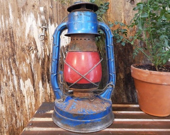 May include: A blue and red vintage oil lamp with a glass globe. The lamp has a metal handle and a metal base. The lamp is sitting on a wooden surface.