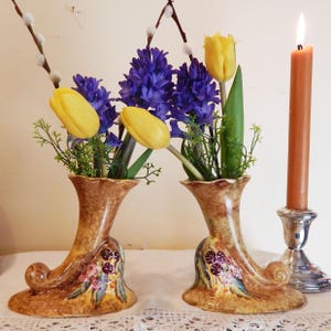 May include: Two brown ceramic vases shaped like a boot with a yellow tulip and purple hyacinth flowers. A lit orange candle is in a silver candlestick.