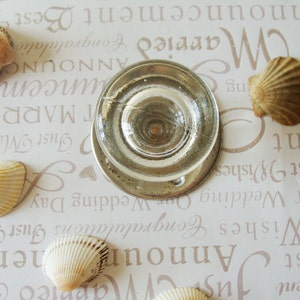 Peut inclure: Un objet en verre transparent de forme circulaire, entouré de plusieurs coquillages. Les coquillages sont beiges et bronzés, et le fond est un papier clair avec du texte imprimé.