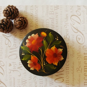 May include: A black, round decorative box with a floral design in red, orange, and yellow. The flowers are painted on the lid with green leaves and stems. Three brown pine cones are arranged near the box. The background is a light-colored card with text.