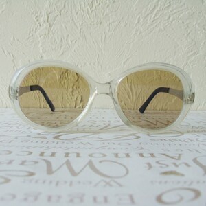 May include: Clear-framed sunglasses with brown-tinted lenses. The sunglasses have a large, rounded shape and black arms. The image is a close-up shot of the sunglasses, showcasing their design and style.
