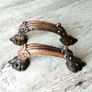 May include: Two antique bronze-colored cabinet handles. Each handle has a curved bar with ornate detailing and decorative end pieces. The handles are designed for easy installation and add a vintage touch to furniture.