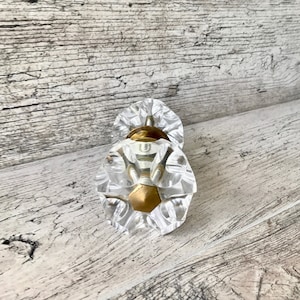 Może przedstawiać: Dwa przezroczyste, fasetowane gałki do szafek ze złotymi metalowymi podstawami. Gałki mają kształt kwiatów i leżą na zwietrzałej, jasnoszarej drewnianej powierzchni. Gałki są przeznaczone do mebli lub dekoracji.