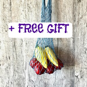 May include: A blue string shopping bag filled with yellow and red produce. The bag is held by a hand against a weathered wood background. The text "+ FREE GIFT" is displayed on a white rectangle.
