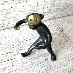 Puede incluir: Pequeña figura de mono negro con cara y detalles de latón. El mono está en una pose juguetona, con un brazo extendido y la cola enroscada. La figura es de metal y está sobre una superficie de madera blanca.