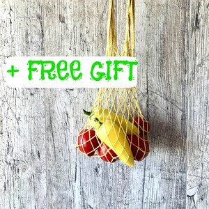 May include: A yellow and beige woven net bag filled with red and yellow bell peppers. The bag is held by a hand against a weathered, light-colored wooden background. The text "+ FREE GIFT" is displayed in green.