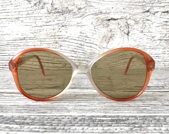 May include: Vintage oversized sunglasses with a gradient lens. The frames are a translucent white with orange accents. The lenses are a light brown color. The sunglasses have a retro style.