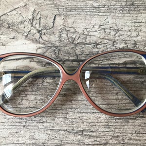 May include: Retro-style eyeglasses. The frames are a combination of peach, blue, and white. The lenses are clear, and the arms are a light green colour. The glasses are set against a textured, light brown wooden surface.