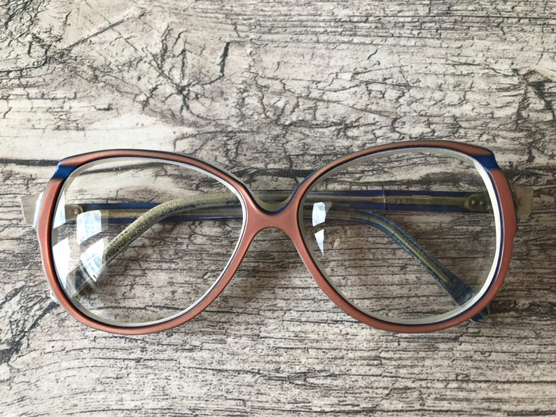 May include: Retro-style eyeglasses. The frames are a combination of peach, blue, and white. The lenses are clear, and the arms are a light green colour. The glasses are set against a textured, light brown wooden surface.