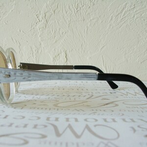 May include: Oversized, clear-framed sunglasses with light brown lenses. The temples are silver with a decorative etched design, transitioning to black earpieces. The glasses are angled, showcasing their retro style.
