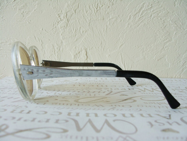 May include: Oversized, clear-framed sunglasses with light brown lenses. The temples are silver with a decorative etched design, transitioning to black earpieces. The glasses are angled, showcasing their retro style.
