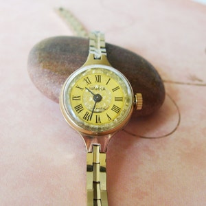 May include: A vintage gold-toned wristwatch with a round, yellow face featuring black Roman numerals and the word "ЧАЙКА". The watch has a gold-toned band and a small crown. The watch is resting on a brown stone.