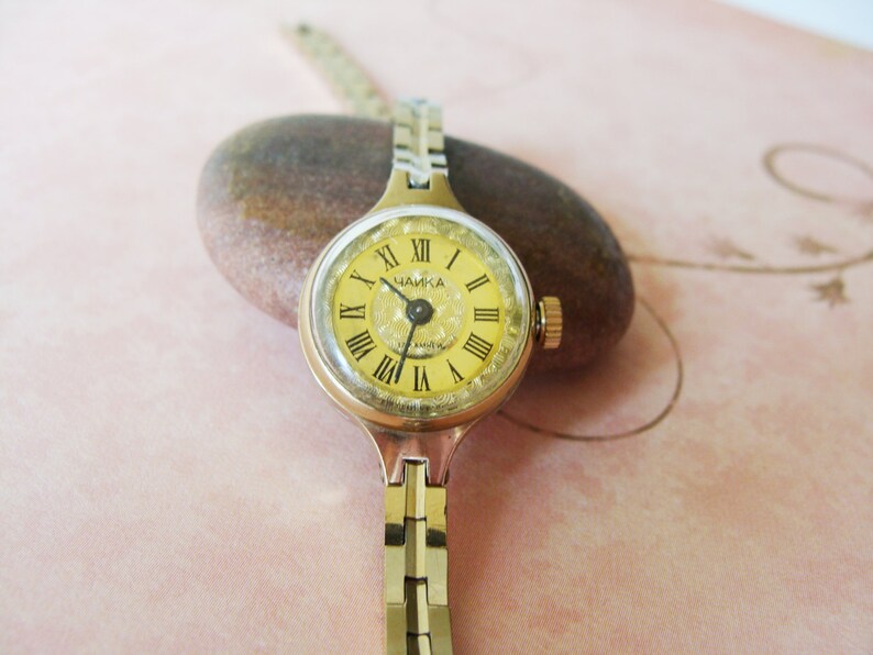May include: A vintage gold-toned wristwatch with a round, yellow face featuring black Roman numerals and the word "ЧАЙКА". The watch has a gold-toned band and a small crown. The watch is resting on a brown stone.
