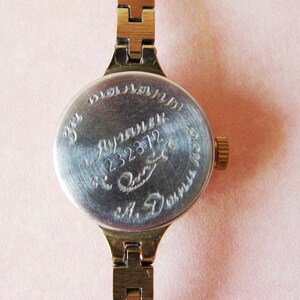 May include: Close-up of a vintage gold-toned wristwatch with a metal band. The watch face is silver with engraved text and numbers. The band is made of rectangular links. The watch is on a pink background.