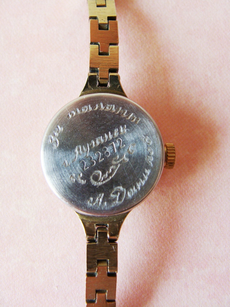 May include: Close-up of a vintage gold-toned wristwatch with a metal band. The watch face is silver with engraved text and numbers. The band is made of rectangular links. The watch is on a pink background.