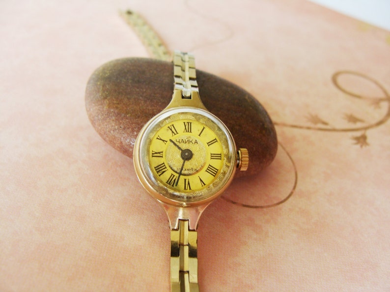 May include: A vintage gold-toned wristwatch with a round, yellow face featuring Roman numerals and the Cyrillic text "ЧАЙКА". The watch has a gold-colored metal band and is resting on a brown stone. The watch face is approximately 1 inch in diameter.