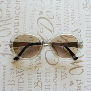 May include: Clear-framed sunglasses with brown lenses. The sunglasses have a rounded shape and black arms. The background is a light-colored surface with wedding-themed text.