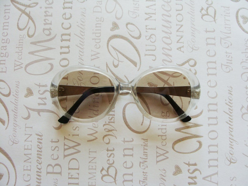 May include: Clear-framed sunglasses with brown lenses. The sunglasses have a rounded shape and black arms. The background is a light-colored surface with wedding-themed text.