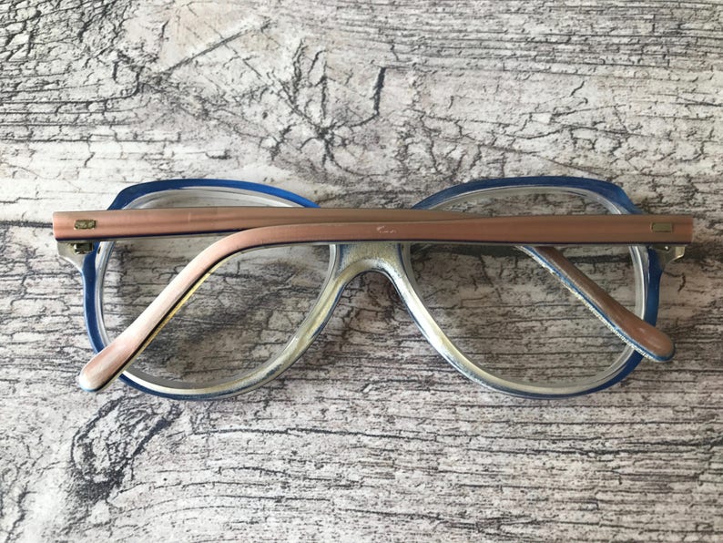 May include: Vintage spectacles with a blue and clear frame and a copper-coloured bridge. The glasses have a retro design, with a rectangular shape and a thick frame. The arms are also copper-coloured.