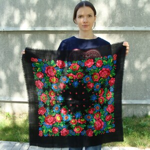 May include: Black square scarf with a floral pattern in red, blue, and yellow. The scarf has a fringed edge and is held up against a gray background. The floral design is centered on the scarf.