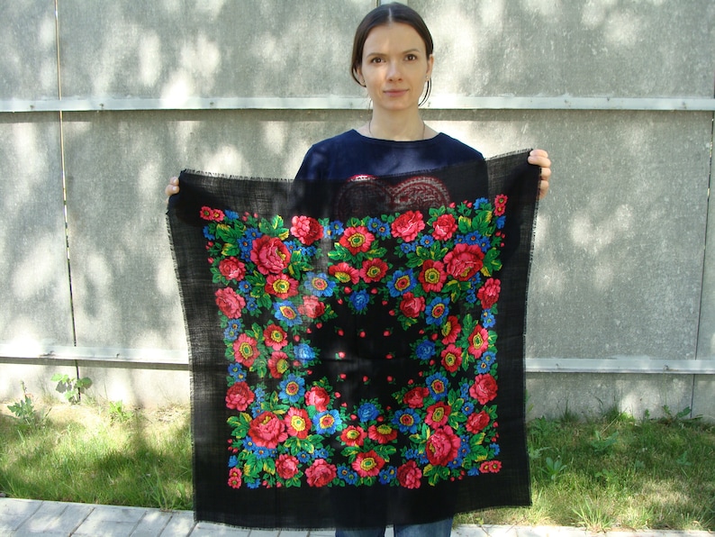 May include: Black square scarf with a floral pattern in red, blue, and yellow. The scarf has a fringed edge and is held up against a gray background. The floral design is centered on the scarf.