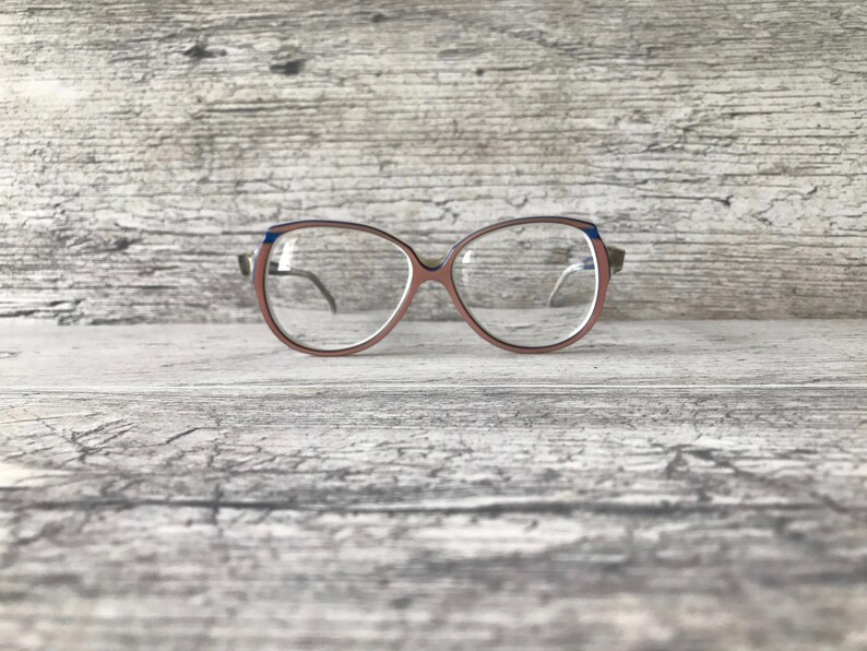May include: Eyeglasses with a light brown frame and blue detailing on the top corners. The lenses are clear, and the arms are a darker brown. The glasses are set against a light grey wooden background.