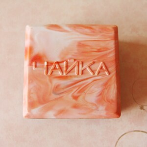 May include: Square soap bar with a swirled peach and white pattern. The word "ЧАЙКА" is embossed on the top. The soap has a marbled appearance, with a mix of light and dark peach tones.