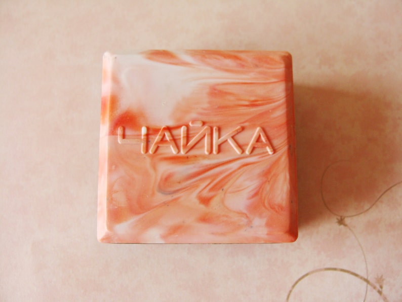 May include: Square soap bar with a swirled peach and white pattern. The word "ЧАЙКА" is embossed on the top. The soap has a marbled appearance, with a mix of light and dark peach tones.