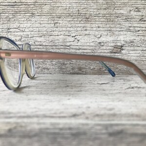 May include: A pair of spectacles with a clear frame and a light brown arm. The frame has a blue accent. The glasses are resting on a light-coloured, textured surface.