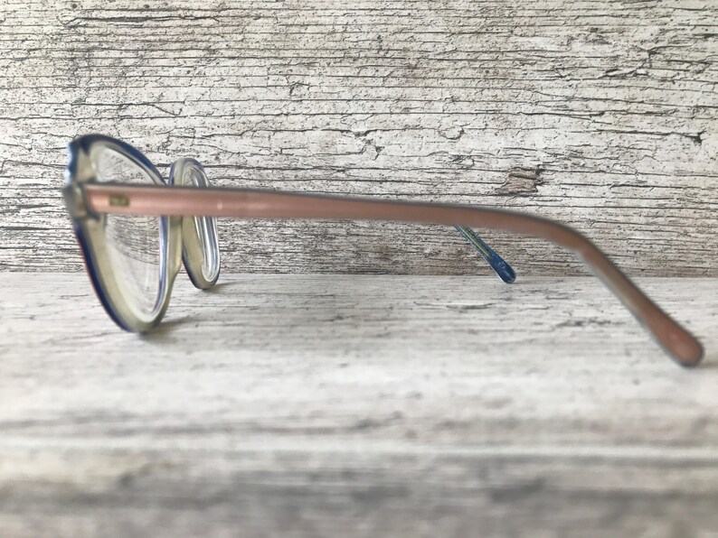May include: A pair of spectacles with a clear frame and a light brown arm. The frame has a blue accent. The glasses are resting on a light-coloured, textured surface.