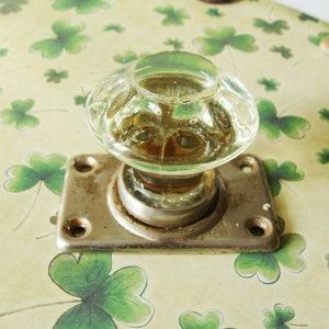 May include: A clear glass doorknob with a decorative, rounded design, mounted on a rectangular, aged metal plate. The knob sits on a background of light green paper with shamrock patterns.