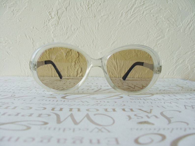 May include: Clear-framed sunglasses with brown-tinted lenses. The sunglasses have a large, rounded shape and black arms. The image is a close-up shot of the sunglasses, showcasing their design and style.