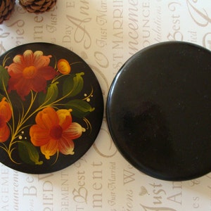 May include: Two round, black wooden coasters. One coaster features a floral design with red, orange, and yellow flowers, green leaves, and gold stems. The other coaster is solid black.