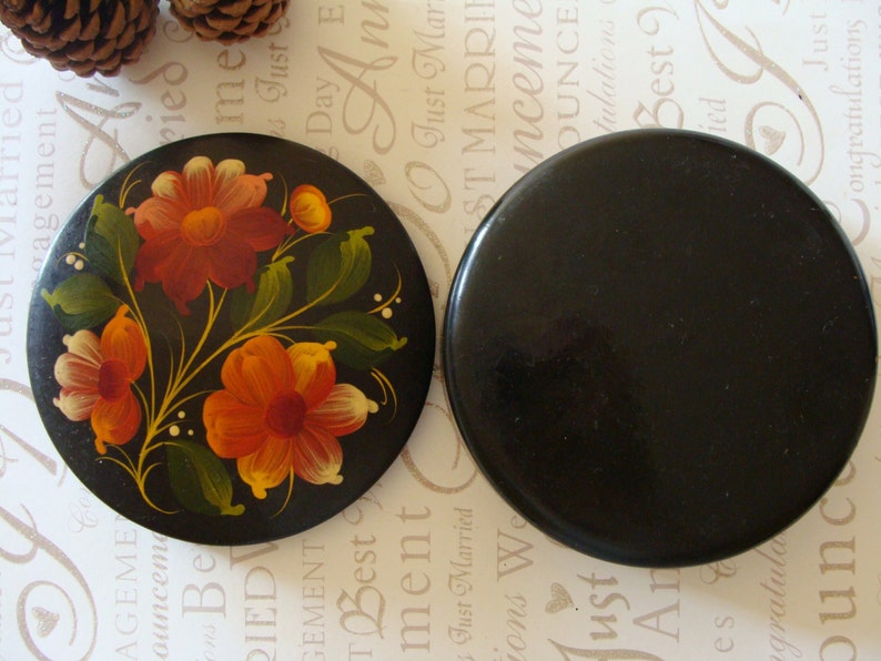 May include: Two round, black wooden coasters. One coaster features a floral design with red, orange, and yellow flowers, green leaves, and gold stems. The other coaster is solid black.